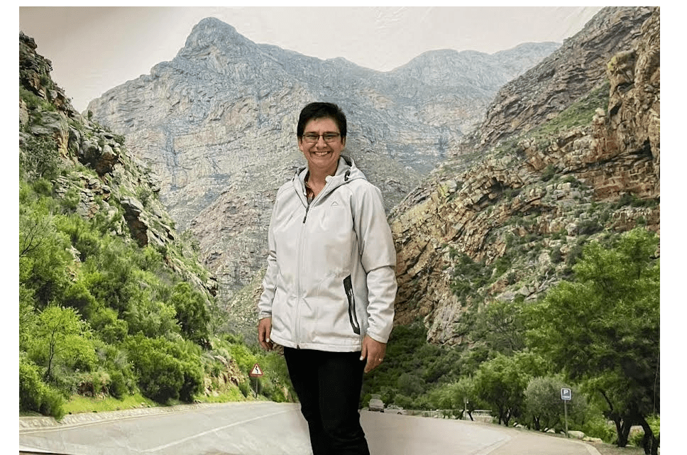 Owner, Elmarie smiling on a scenic mountain road surrounded by rocky cliffs and lush greenery – outdoor portrait with natural South African landscape backdrop.
